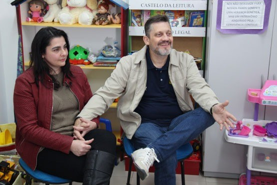 Foto mostra o casal que participa do serviço de família acolhedora, sentados e conversando em uma sala com brinquedos infantis.