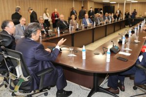 Foto mostra momento da reunião, com participantes sentados em longa mesa em U. Em destaque, de costas, o ministro Fux falando.