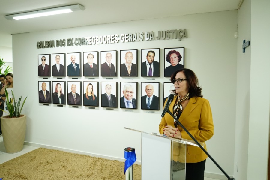 Foto mostra a corregedora-geral do TJTO discursando em um púlpito em frente à galeria de fotos inaugurada.