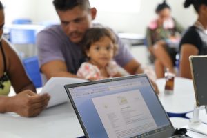 Foto mostra, em primeiro plano, um computador conectado a um celular. Ao fundo, sentados e sendo atendidos, um homem, uma mulher e uma criança.