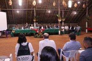 Foto mostra momento da cerimônia de lançamento dentro de uma grande maloca de sapê/palha. Na foto, vê-se a mesa diretiva do evento, com diversas pessoas sentadas.