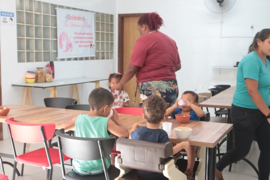 Foto mostra refeitório de unidade de acolhimento. Em uma mesa, três crianças estão sentadas e se alimentando. Ainda há, ao fundo, uma cuidadora em pé, dando um copo de leite para uma menina.