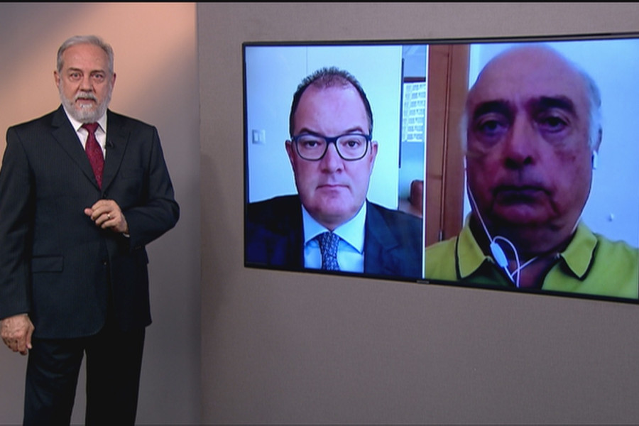 Foto no estúdio do programa mostra o apresentador posando em pé ao lado da telão, onde estão os rostos dos dois entrevistados da edição.