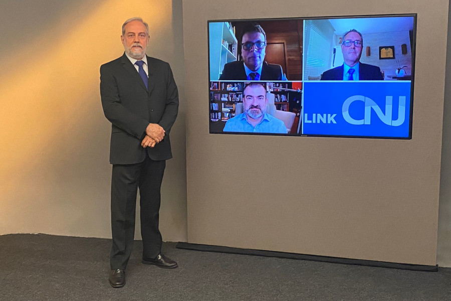 Foto mostra o apresentador no estúdio, posando ao lado do telão onde se vê os três entrevistados do programa participando por videoconferência.