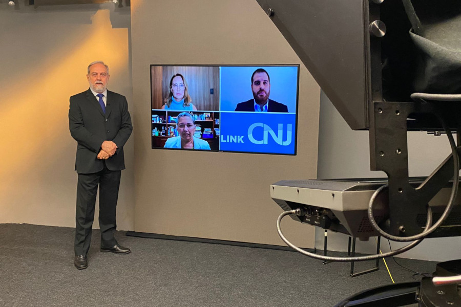 Foto do estúdio do programa com o apresentador posando ao lado do telão onde se vê os entrevistados por videoconferência.