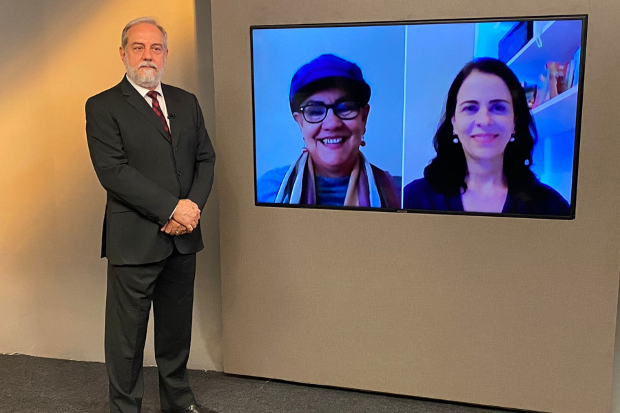 Foto do estúdio do programa com o apresentador posando ao lado do telão onde se vê as entrevistados por videoconferência.