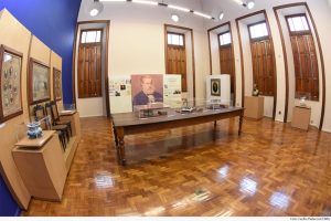 Foto mostra espaço de exposição no Memória do Judiciário Mineiro., onde se vê uma mesa ao centro com objetos expostos em redomas, quadros nas paredes e paineis.