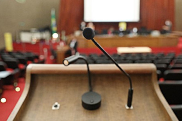 Foto mostra em destaque um púlpito com um microfone e, ao fundo, o auditório e o palco do Plenário do TSE.