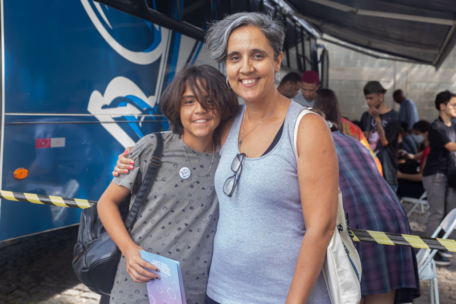 Foto mostra mãe e filho abraçados em frente ao ônibus do projeto e diversas pessoas que estão sendo atendidas em mesas do lado de fora do ônibus.