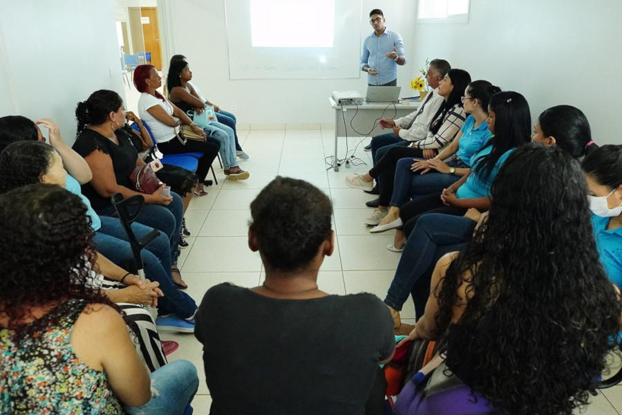 Foto mostra sala do Escritório Social, com as pessoas sentadas em cadeiras, em formato de U. Ao fundo, um homem em pé está fazendo uma apresentação, que está sendo exibida na parede com um datashow.