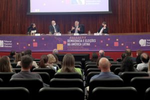 Foto mostra o palco visto do fundo do auditório do TSE. No palco, sentados atrás de uma mesa, estão o palestrante, o ministro Edson Fachin e mais duas pessoas.