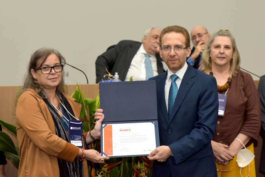 Foto mostra momento da entrega da menção honrosa, com uma mulher recebendo o certificado das mãos de um homem.