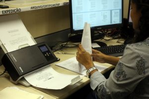 Foto mostra uma mulher, em uma estação de trabalho com computador, escaneando folhas de processos.