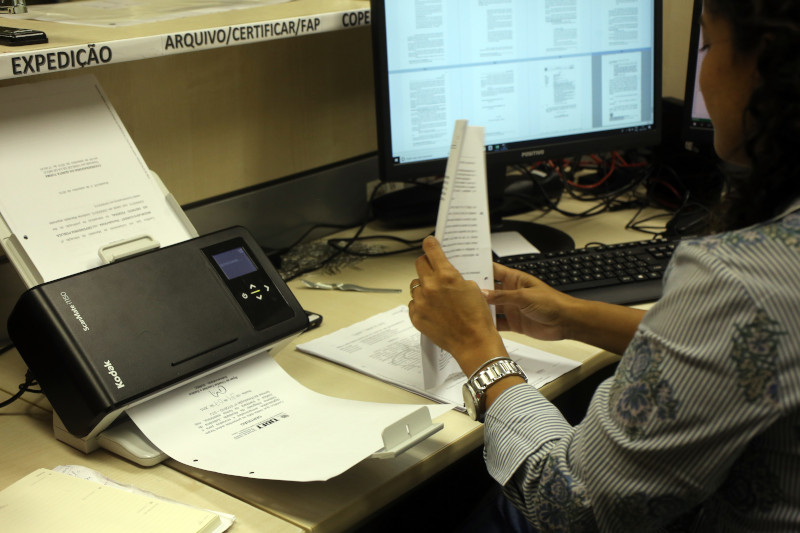 Foto mostra uma mulher, em uma estação de trabalho com computador, escaneando folhas de processos.
