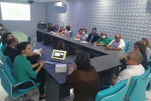 Foto mostra momento da reunião em uma sala do presídio, com as pessoas sentadas em uma mesa em U e, ao fundo, com uma tela de apresentação sendo exibida na parede por um datashow.