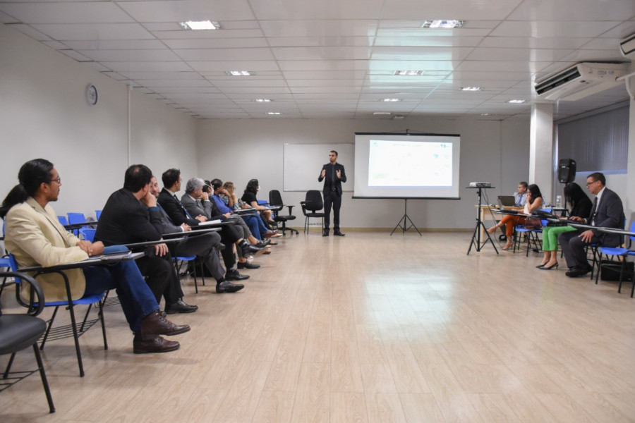 Foto mostra pessoas em uma sala, assistindo a uma apresentação de um homem que ao lado de um telão.