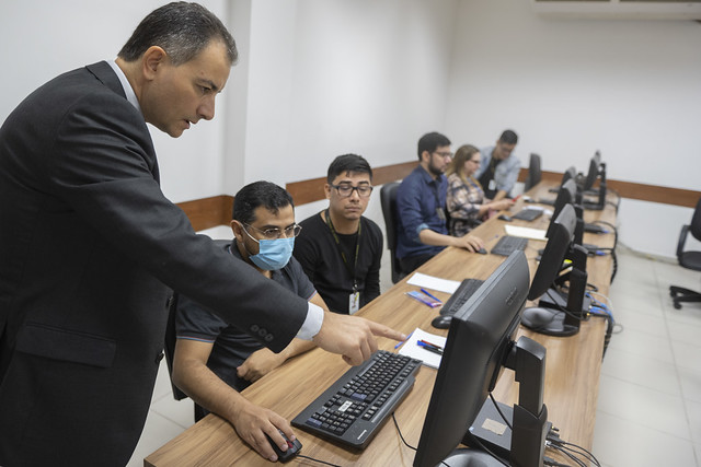 Foto mostra cinco pessoas em uma bancada com computadores participando do treinamento e, em primeiro plano, um homem em pé orientando um dos alunos.