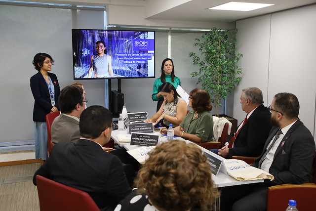 Foto mostra, em uma sala, oito pessoas sentadas em volta de uma mesa, duas mulheres em pé à frente e, entre elas, tem um telão onde se lê "Protocolo de Escuta Qualificada para Grupos Vulneráveis ao Tráfico de Pessoas".