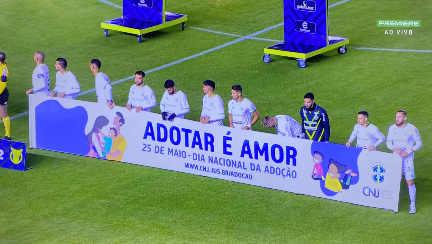 Foto de time de futebol, antes do início de partida, segurando a faixa da campanha Adotar É Amor.