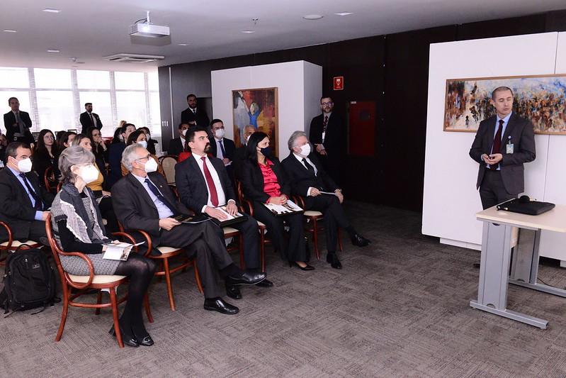 Foto mostra momento da apresentação, com cerca de 20 pessoas sentadas e uma à frente falando.