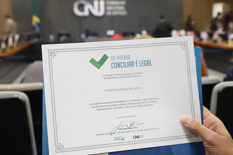Foto mostra um certificado da premiação e, ao fundo, desfocado, o Plenário do CNJ durante o evento.