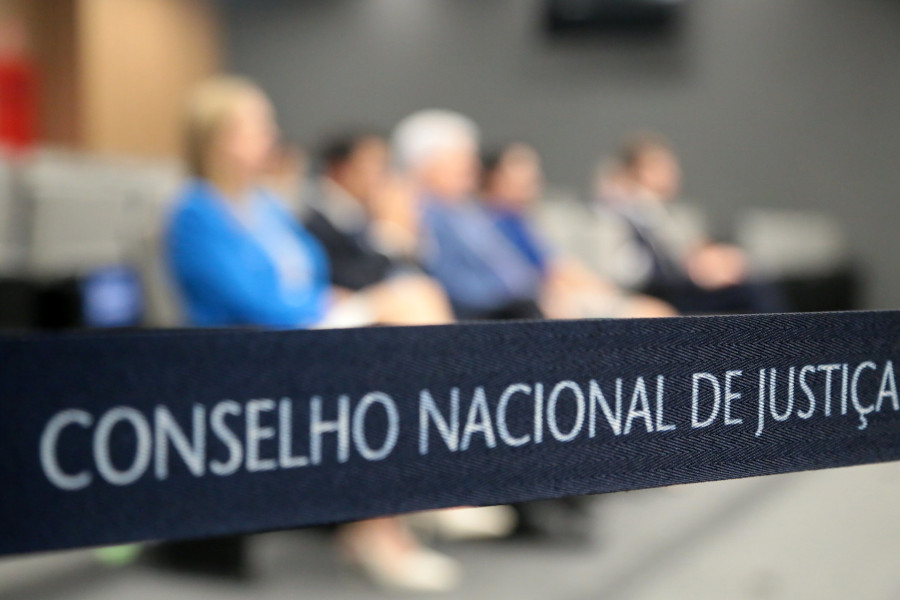Foto mostra divisor de fila com o nome do Conselho Nacional de Justiça e, ao fundo e desfocado, é possível ver cinco pessoas sentadas no Plenário do CNJ e acompanhando a sessão.