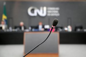 Foto mostra visão frontal do Plenário do CNJ, estando ao fundo, desfocados, conselheiros que compõem a bancada principal, com a logomarca do CNJ na parede. Em primeiro plano, o foco está no microfone onde são feitas as participações de representantes e partes.