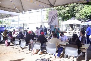 Foto mostra uma mesa comprida, montada sob uma grande tenda, com pessoas sentadas e sendo atendidas.