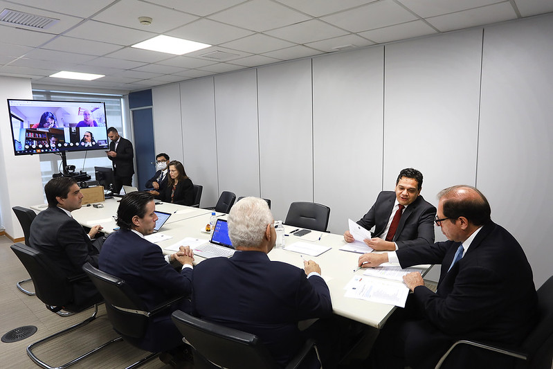 Foto mostra uma sala de reuniões, com pessoas sentadas em volta de uma mesa conversando e um telão ao fundo com participantes por videoconferência.