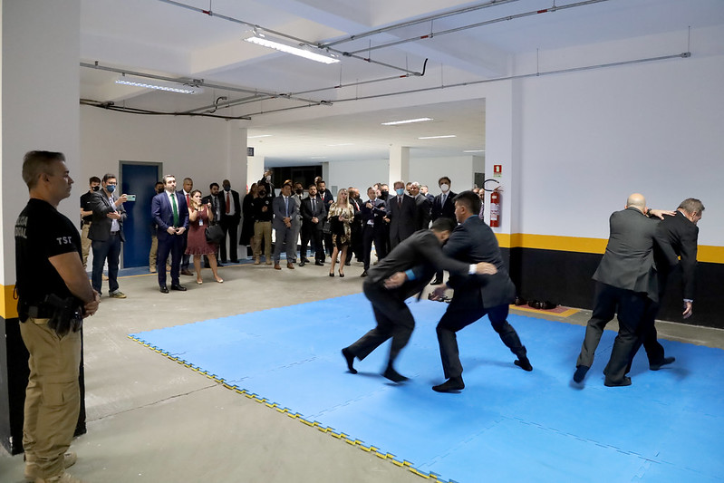 Foto mostra quatro homens em um tatame demonstrando técnicas de defesa pessoal enquanto as pessoas participantes do evento assistem em volta.