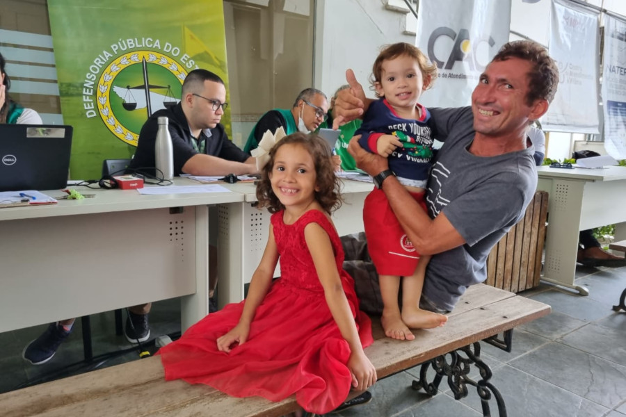 Foto de André Nascimento e seus dois filhos sentados em um banco e sorrindo para a fotografia enquanto aguardam para serem atendidos pela Defensoria Pública do estado.
