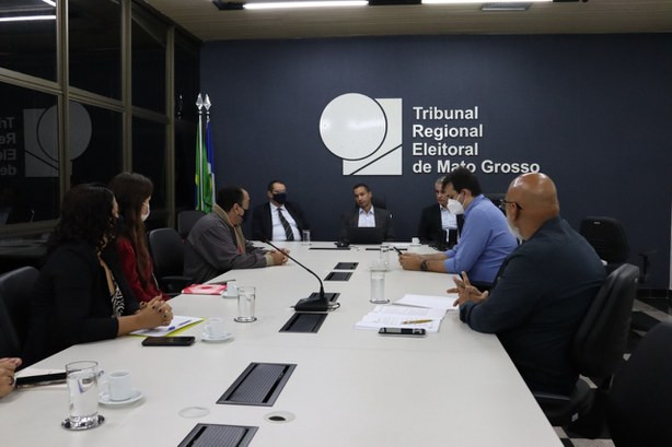 Foto da sala de reuniões do TRE-MT, que tem uma mesa comprida, onde aparecem os participantes sentados ao redor. No fundo, tem uma parede azul com a marca do TRE-MT na cor branca.