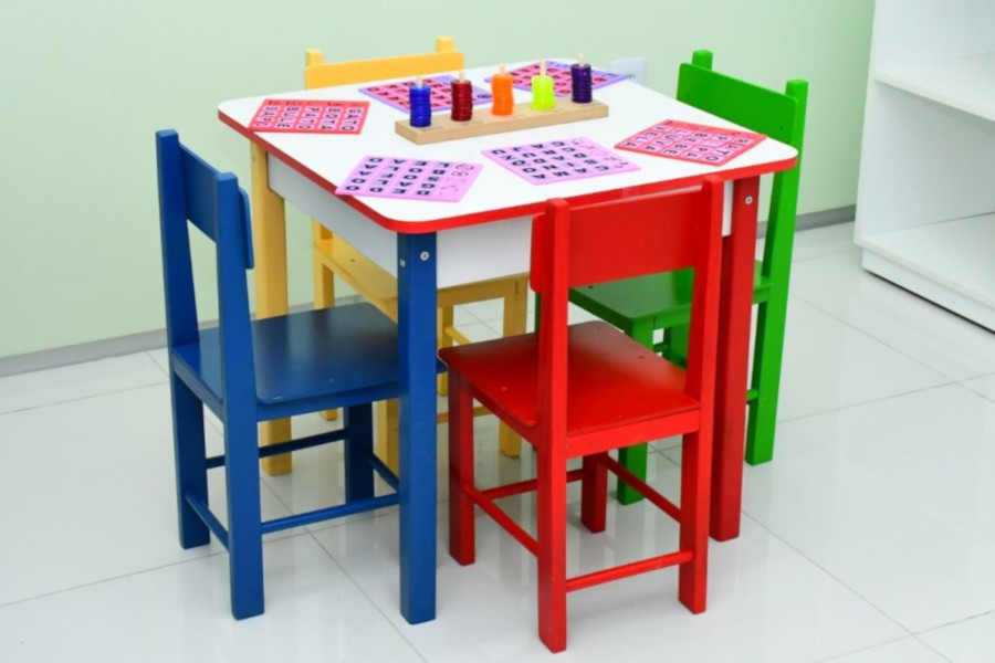 Foto mostra sala com uma mesa infantil e quatro cadeiras coloridas ao centro e alguns brinquedos em cima dela.