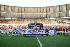 Foto mostra jogadores de time de futebol, em um estádio cheio, enfileirados antes do jogo para ouvir o hino nacional. Na frente deles, uma faixa da campanha.