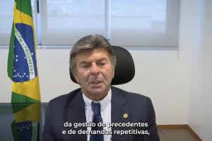 Foto do ministro Luiz Fux fazendo seu discurso na abertura do evento. Ele participa por videoconferência e está sentado em uma sala, com a bandeira do Brasil ao fundo.