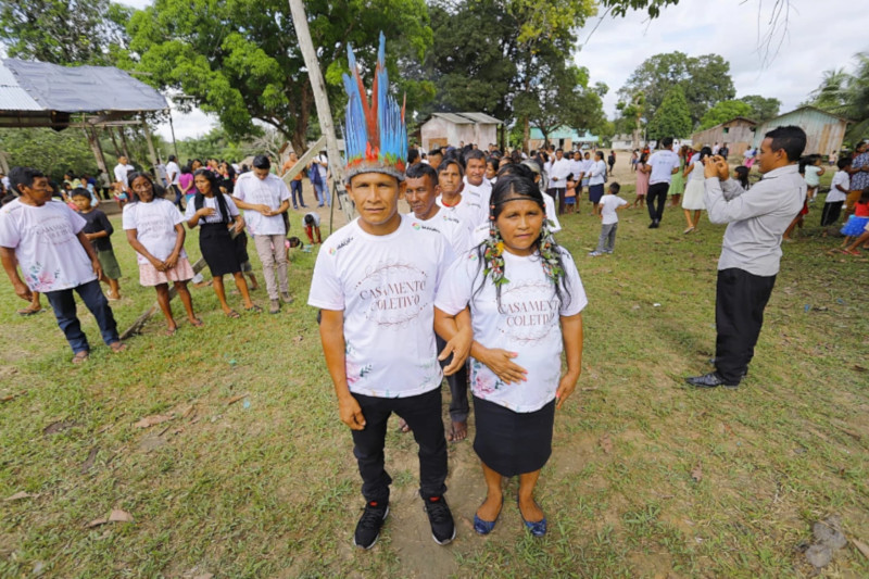 Foto mostra um casal de homem e mulher indígenas e, atrás deles, uma fila com outros casais. Eles estão em um espaço aberto, com grama e árvores e diversas pessoas assistindo.