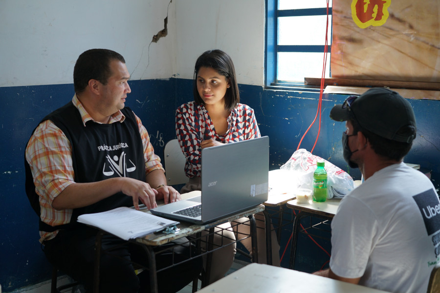 Foto mostra o professor e uma aluna sentados em carteiras escolares e com um notebook à frente. Eles estão atendendo a um homem que está sentado na frente deles.