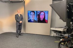 Foto do estúdio do programa com o apresentador posando ao lado do telão onde se vê as pessoas entrevistadas por videoconferência.