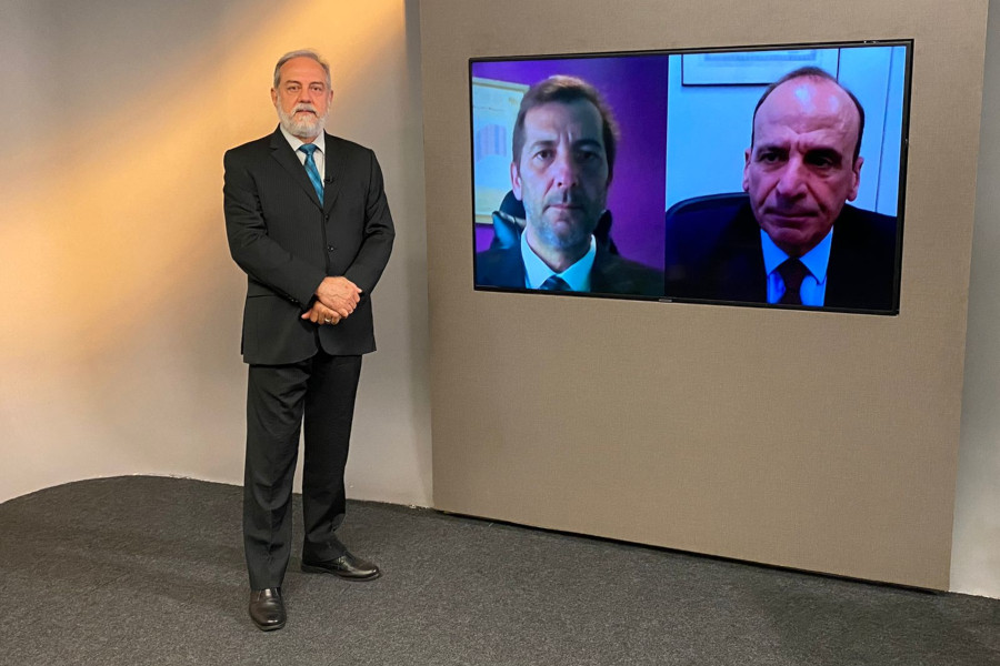 Foto do estúdio do programa com o apresentador posando ao lado do telão onde se vê as pessoas entrevistadas por videoconferência.