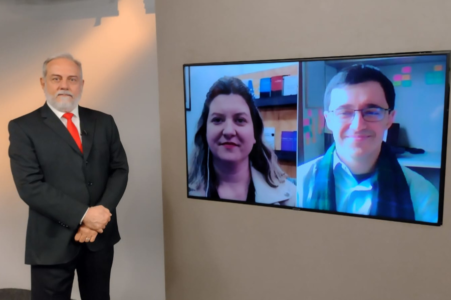 Foto do estúdio do programa com o apresentador posando ao lado do telão onde se vê as pessoas entrevistadas por videoconferência.