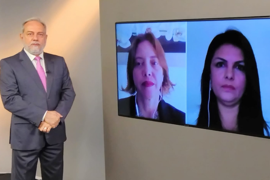 Foto do estúdio do programa com o apresentador posando ao lado do telão onde se vê as pessoas entrevistadas por videoconferência.