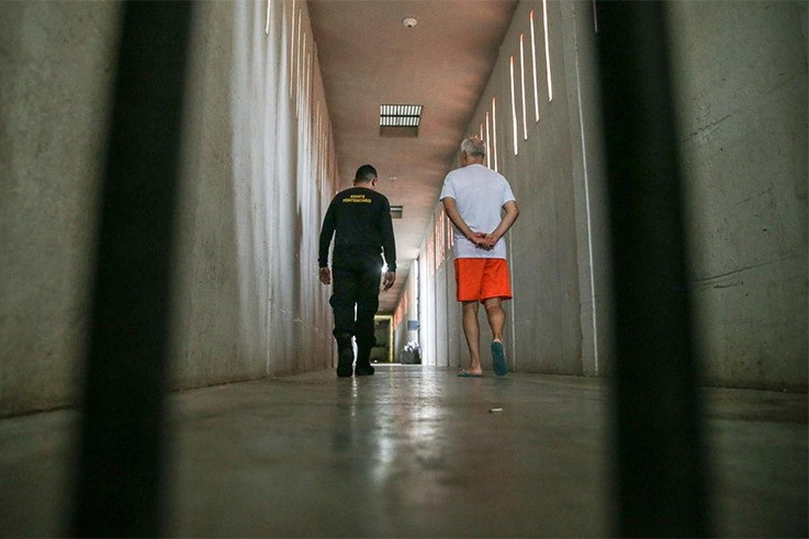 Foto mostra em um corredor de presídio, atrás de grades, um policial conduzindo um detento.