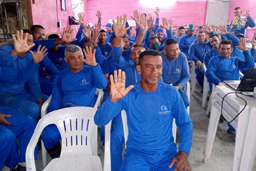 Foto mostra os homens, trabalhadores da construção civil usando camisa e calça azul, sentados em uma sala e mostrando a palma da mão com um X vermelho.