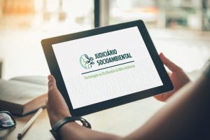 Foto mostra uma mulher segurando um tablet. Na tela, tem a logomarca do evento "Judiciário Socioambiental".