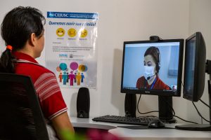 Foto mostra uma mulher de costas. sentada à frente do computador pelo qual participa de uma audiência virtual. Ela esta em uma sala física do Centro Judiciário de Solução de Conflitos e Cidadania de Cuiabá.