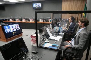 Foto mostra dois homens no Plenário do CNJ assistindo aos demais participantes do evento por um telão central onde se vê a videoconferência.