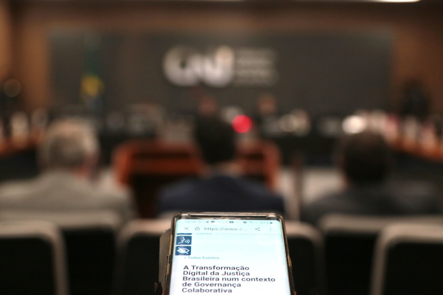 Foto mostra uma visão geral do plenário do CNJ durante o evento, desfocada. Em primeiro plano, um celular com o título do evento.