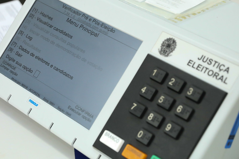 Foto mostra uma urna eletrônica com o menu "Verificador Pré e Pós Eleição" na tela.