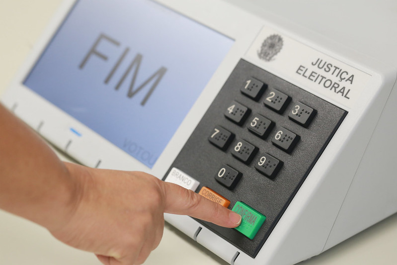 Foto mostra a mão de um homem apertando a tecla Confirma em uma urna eletrônica. Na tela, tem o texto "FIM".