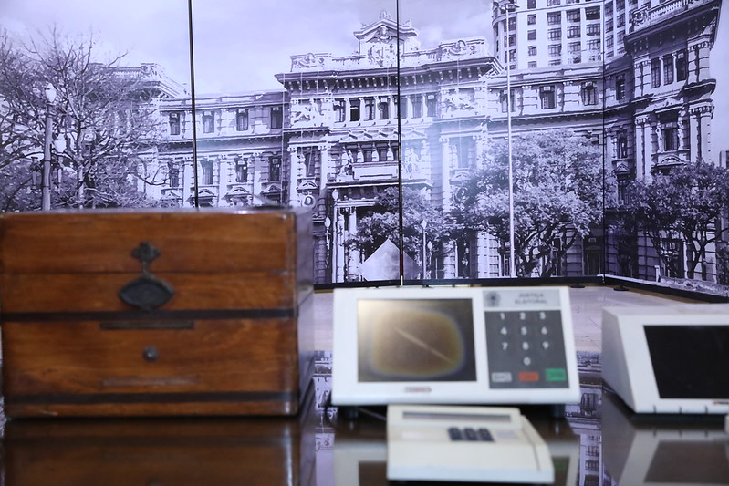 Foto mostra, em primeiro plano, urnas eleitorais em madeira e eletrônica desfocadas. Ao fundo, painel com a foto do Palácio da Justiça de São Paulo.